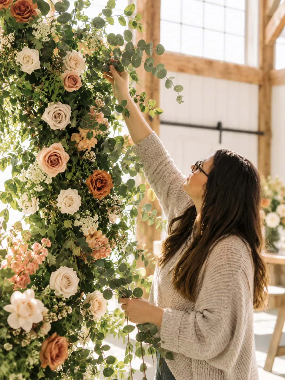 A florist reaches up to place eucalyptus stems on a tall installation of roses and greenery inside a sunlit, timber-framed venue.